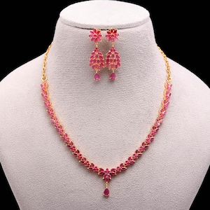 Op de afbeelding: Een goudkleurige ketting- en oorbellenset. De ketting en oorbellen zijn versierd met roze edelstenen. De ketting heeft een druppelvormige hanger. De oorbellen hebben een bloemenontwerp en een waterval van edelstenen.