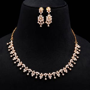 May include: A gold-toned necklace and matching earrings set. The jewelry features a floral design with small, clear gemstones. The necklace curves gracefully, and the earrings are composed of multiple floral clusters. The set is displayed on a black background.