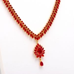 May include: A gold necklace adorned with red gemstones. The chain is set with small, leaf-like red stones, culminating in a flower-shaped pendant. A teardrop-shaped stone hangs below. The necklace is presented on a white backdrop, highlighting the jewellery's design.