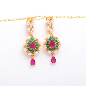 May include: A pair of gold-toned earrings with a teardrop design. Each earring features a central pink gemstone surrounded by green and white stones. A pink teardrop hangs below the central design. The earrings are suspended from a gold chain.