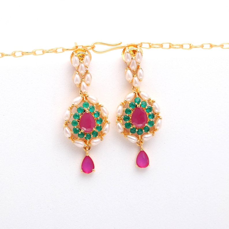 May include: A pair of gold-toned earrings with a teardrop design. Each earring features a central pink gemstone surrounded by green and white stones. A pink teardrop hangs below the central design. The earrings are suspended from a gold chain.