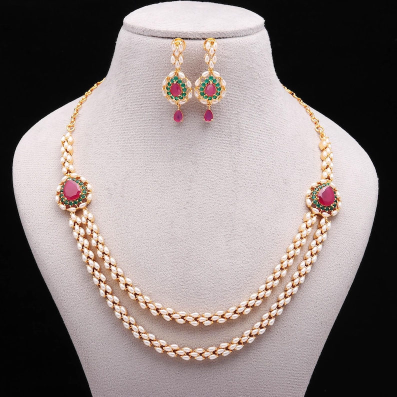 May include: A two-strand pearl necklace and matching earrings set. The necklace features a large, oval, red gemstone surrounded by small green stones. The earrings have a similar design, with a red teardrop-shaped gemstone hanging from each earring.