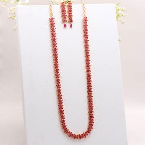 May include: A gold necklace and earring set featuring a repeating pattern of red gemstones. The necklace is long and hangs vertically. The matching earrings have a similar design. The jewellery is displayed on a white surface.