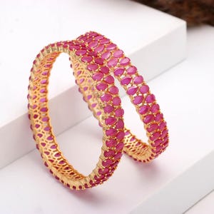Può includere: Due braccialetti dorati con un motivo ripetuto di pietre rosa a forma di goccia. I braccialetti sono esposti su una superficie bianca, evidenziando il design intricato e il colore vibrante dei gioielli.
