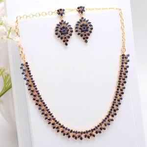 May include: A gold-toned jewelry set featuring a necklace and matching earrings. The necklace and earrings are adorned with numerous dark blue gemstones. The earrings have a teardrop shape. The jewelry is displayed on a white surface.