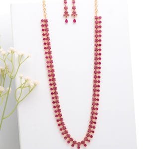 May include: A gold-toned necklace and matching earrings set. The necklace and earrings are adorned with small, round, and teardrop-shaped red gemstones. The jewelry is displayed on a white surface, with a sprig of white flowers in the background.