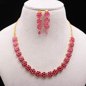 May include: A gold-coloured necklace and matching earrings set. The necklace and earrings feature a floral design with vibrant pink gemstones. The earrings are drop style, and the necklace is a choker style.