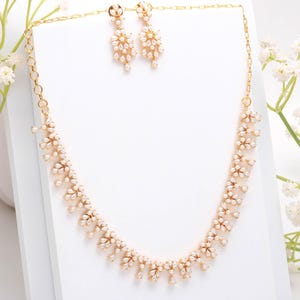 May include: A gold-toned jewelry set featuring a necklace and matching earrings. The necklace and earrings are adorned with small, white, pearl-like accents arranged in a floral pattern. The set is displayed on a white surface.