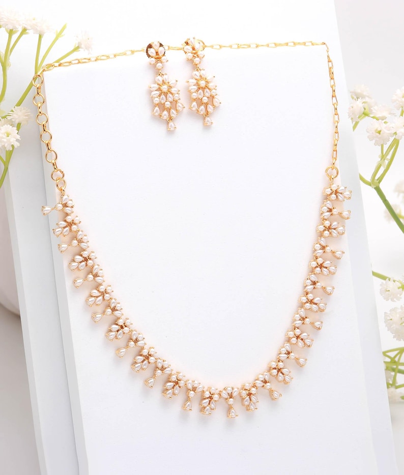 May include: A gold-toned jewelry set featuring a necklace and matching earrings. The necklace and earrings are adorned with small, white, pearl-like accents arranged in a floral pattern. The set is displayed on a white surface.