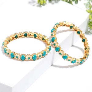 Pode incluir: Duas pulseiras douradas com pedras ovais turquesa e pequenos detalhes brancos. As pulseiras s&atilde;o exibidas em uma superf&iacute;cie branca, destacando o design e o contraste de cores das joias. As pedras turquesas s&atilde;o espa&ccedil;adas uniformemente.