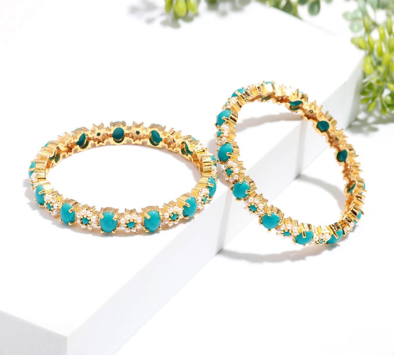 Pode incluir: Duas pulseiras douradas com pedras ovais turquesa e pequenos detalhes brancos. As pulseiras s&atilde;o exibidas em uma superf&iacute;cie branca, destacando o design e o contraste de cores das joias. As pedras turquesas s&atilde;o espa&ccedil;adas uniformemente.