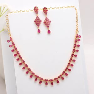 May include: A gold-toned jewelry set featuring a necklace and matching earrings. The necklace and earrings are adorned with vibrant, teardrop-shaped, ruby-red gemstones. The earrings dangle elegantly, and the necklace gracefully curves.