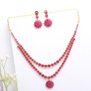 Puede incluir: Un conjunto de collar y pendientes dorados con piedras preciosas rosas vibrantes. El collar tiene un diseño de doble cadena con un colgante, y los pendientes tienen un diseño floral a juego. La joyería está sobre un fondo blanco.