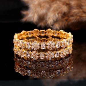 Op de afbeelding: Twee goudkleurige armbanden met een bloemen- en rechthoekig edelsteenontwerp. De armbanden worden weerspiegeld op een donkere ondergrond, waardoor een symmetrisch beeld ontstaat. De heldere edelstenen schitteren, wat bijdraagt aan de elegantie.