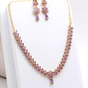 May include: A gold-toned necklace and earring set. The necklace features a leaf-like design with purple gemstones. The earrings have a floral design with a teardrop-shaped purple gemstone. The jewelry is displayed on a white surface.