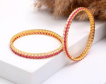 Könnte beinhalten: Zwei goldene Armreifen mit abwechselnd rosa und klaren Edelsteinen. Die Armreifen liegen auf einer weißen Oberfläche, mit einem verschwommenen Hintergrund aus getrockneten Pflanzen. Der Schmuck ist mit einem sich wiederholenden Edelsteinmuster versehen.