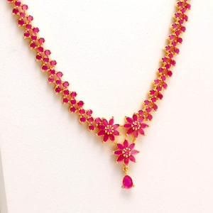 Puede incluir: Un collar dorado con un diseño floral con piedras preciosas rosas vibrantes. El collar tiene una delicada cadena con grupos de piedras rosas y colgantes en forma de flor. Una piedra rosa en forma de lágrima cuelga del centro.