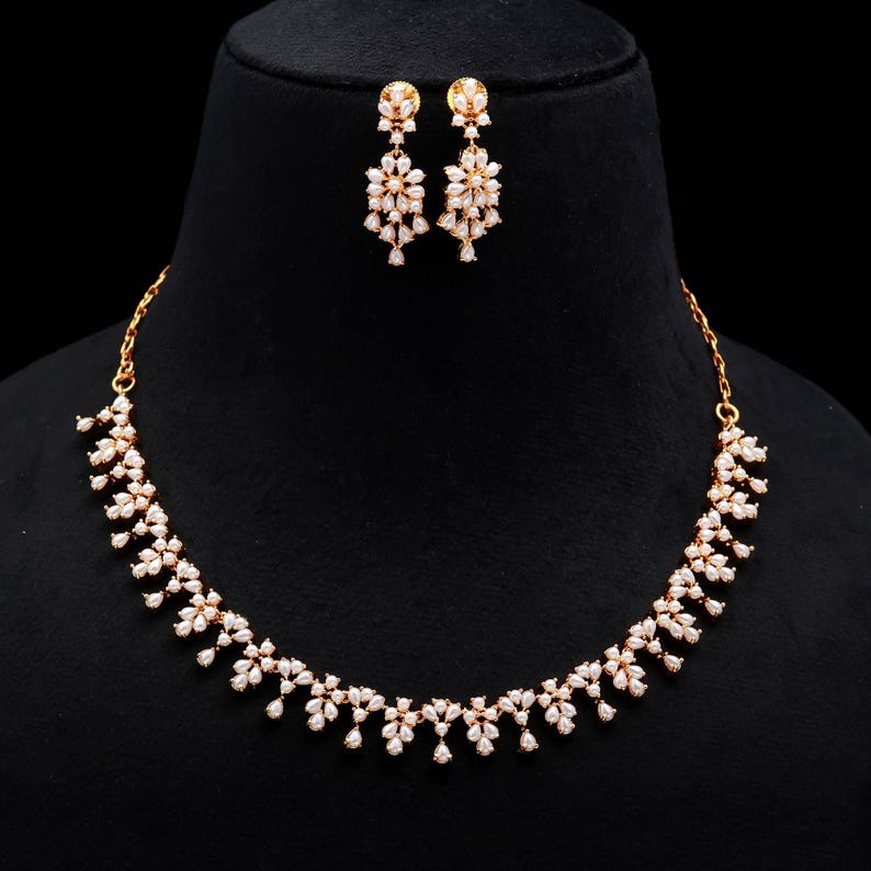 May include: A gold-toned necklace and matching earrings set. The jewelry features a floral design with small, clear gemstones. The necklace curves gracefully, and the earrings are composed of multiple floral clusters. The set is displayed on a black background.