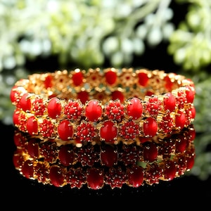 Könnte beinhalten: Zwei goldfarbene Armreifen mit leuchtend roten, ovalen Steinen und kleineren roten Blumenakzenten. Die Armreifen sind gestapelt und spiegeln sich auf einer dunklen Oberfläche wider, was das aufwendige Design und die Handwerkskunst des Schmucks zeigt.