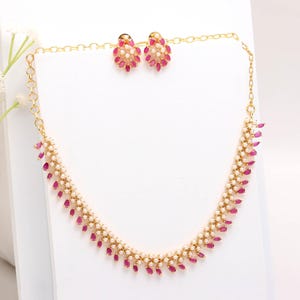 Könnte beinhalten: Ein goldfarbenes Halsketten- und Ohrring-Set. Die Halskette und die Ohrringe weisen ein Muster aus kleinen, weißen Perlen und leuchtend rubinroten, tropfenförmigen Steinen auf. Der Schmuck wird auf einer weißen Oberfläche präsentiert.