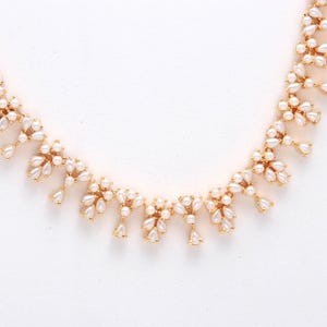 May include: A gold-toned necklace featuring clusters of small, white, pear-shaped faux pearls. The necklace has a delicate design with the pearls arranged in a floral pattern, creating a sophisticated and elegant look. The necklace is set against a white background.