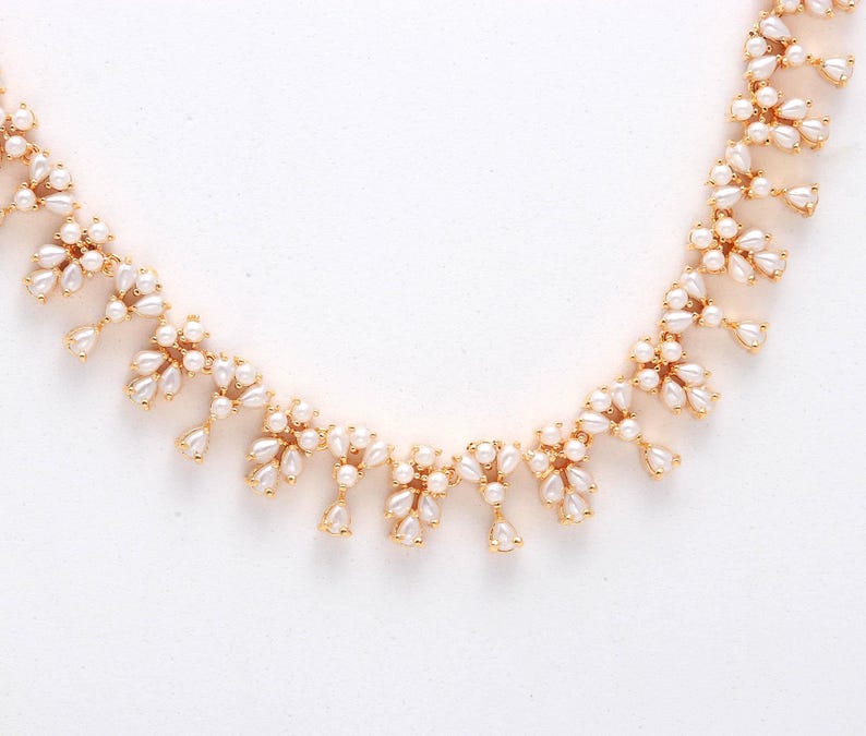 May include: A gold-toned necklace featuring clusters of small, white, pear-shaped faux pearls. The necklace has a delicate design with the pearls arranged in a floral pattern, creating a sophisticated and elegant look. The necklace is set against a white background.