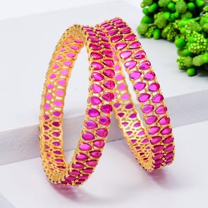 May include: Two gold bangles with a repeating pattern of pink teardrop-shaped stones. The bangles are displayed on a white surface, with green foliage in the background. The bangles are a jewelry item.