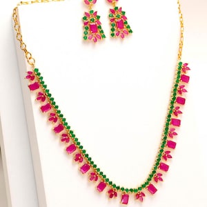 May include: A gold-toned necklace and earring set. The necklace and earrings feature a pattern of alternating fuchsia-colored rectangular stones and green circular stones. The earrings are dangling, and the necklace is a choker style.