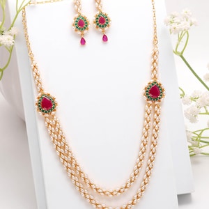 May include: A gold-toned jewelry set featuring a necklace and matching earrings. The necklace has two strands of small, white beads with a central pendant and two side pendants, each with a red gemstone surrounded by green stones. The earrings have a similar design.