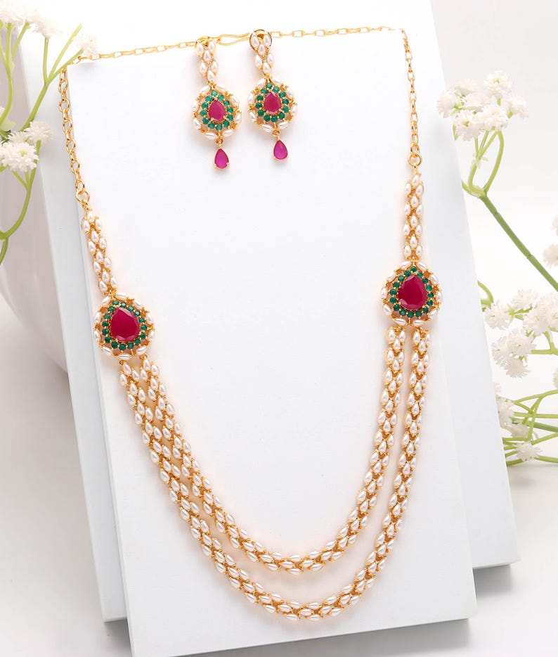 May include: A gold-toned jewelry set featuring a necklace and matching earrings. The necklace has two strands of small, white beads with a central pendant and two side pendants, each with a red gemstone surrounded by green stones. The earrings have a similar design.