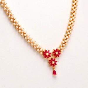 May include: A gold necklace with a double strand of small, white pearls. The necklace features three red flower pendants with white centres and a single teardrop-shaped red pendant. The necklace is set against a white backdrop.