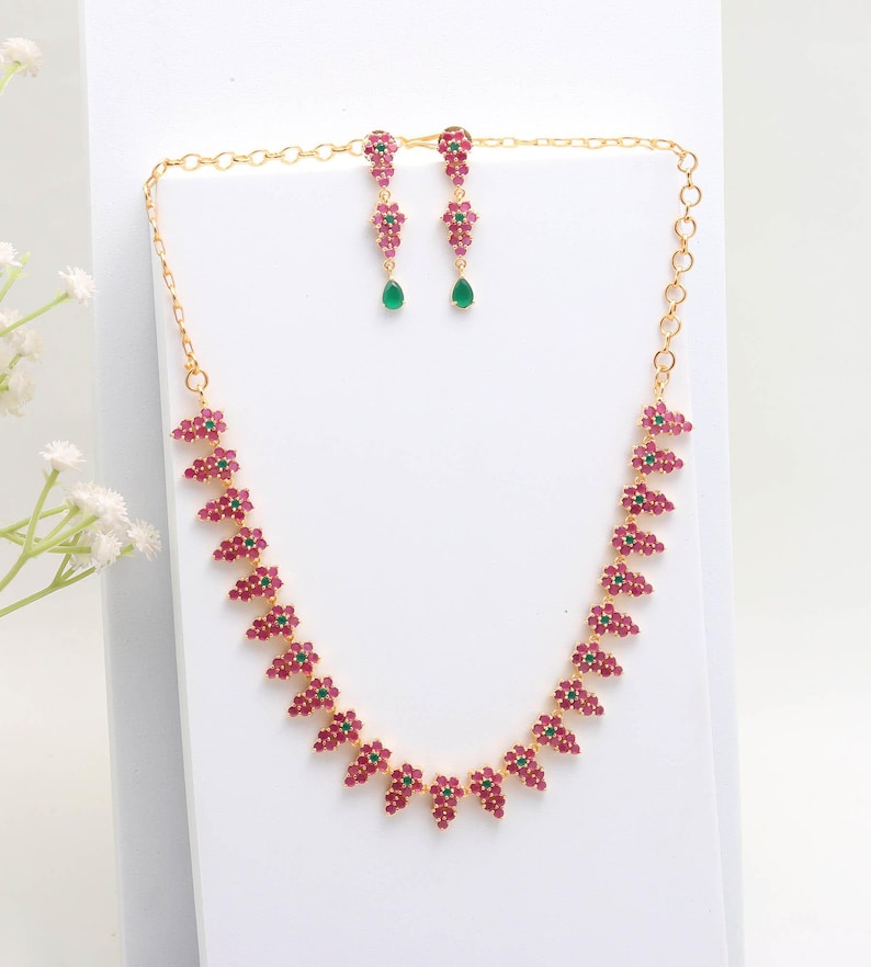 May include: A gold-toned necklace and earring set. The necklace and earrings feature clusters of small, red gemstones interspersed with green gemstones. The earrings have a teardrop-shaped green gemstone at the bottom.