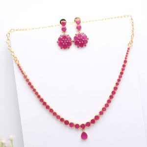 Pode incluir: Um conjunto de colar e brincos a condizer em tom dourado. O colar e os brincos apresentam pedras preciosas rosa vibrantes. Os brincos têm um design floral e o colar tem um pendente em forma de gota. As joias são exibidas numa superfície branca.