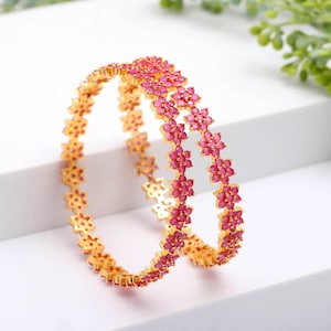 Może przedstawiać: Dwie złote bransoletki z kwiatowym wzorem. Każdy kwiat jest ozdobiony małymi, różowymi kamieniami szlachetnymi. Bransoletki są prezentowane na białej powierzchni, ukazując ich skomplikowane detale i żywy kolor.