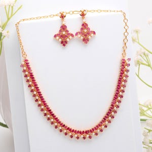 May include: A gold-coloured jewellery set comprising a necklace and matching earrings. The necklace and earrings are decorated with ruby-coloured stones and small white pearls. The earrings are diamond-shaped.