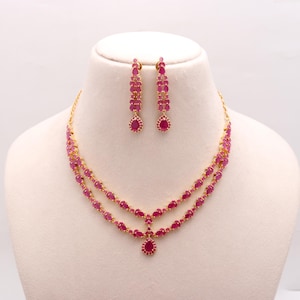 May include: A gold necklace and earring set with ruby-coloured gemstones. The necklace has a double-strand design with a central pendant. The earrings are long, dangling, and also feature ruby-coloured stones. The jewellery is displayed on a white mannequin.