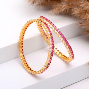 Può includere: Due braccialetti dorati con file alternate di pietre preziose rosa e trasparenti. I braccialetti sono presentati su una superficie bianca, con uno sfondo neutro. I gioielli sono di design classico.