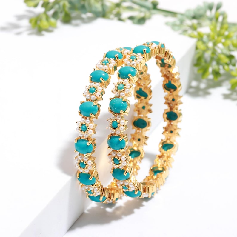 Pode incluir: Duas pulseiras douradas com pedras ovais turquesa e pequenos detalhes de p&eacute;rolas brancas. As pulseiras est&atilde;o dispostas lado a lado, mostrando seu design intrincado. As joias s&atilde;o apresentadas em um fundo branco.