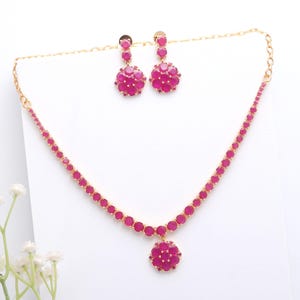 Puede incluir: Un conjunto de joyas de color dorado con un collar y pendientes a juego. El collar y los pendientes están adornados con piedras preciosas redondas y fucsias brillantes. El collar tiene un colgante floral y los pendientes tienen un diseño de racimo.