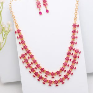 Könnte beinhalten: Ein goldfarbenes Halsketten- und Ohrring-Set. Die Halskette hat zwei Stränge aus rubinroten floralen Designs. Die Ohrringe sind passend dazu mit rubinroten Tropfenanhängern gestaltet. Der Schmuck wird auf einer weißen Oberfläche präsentiert.