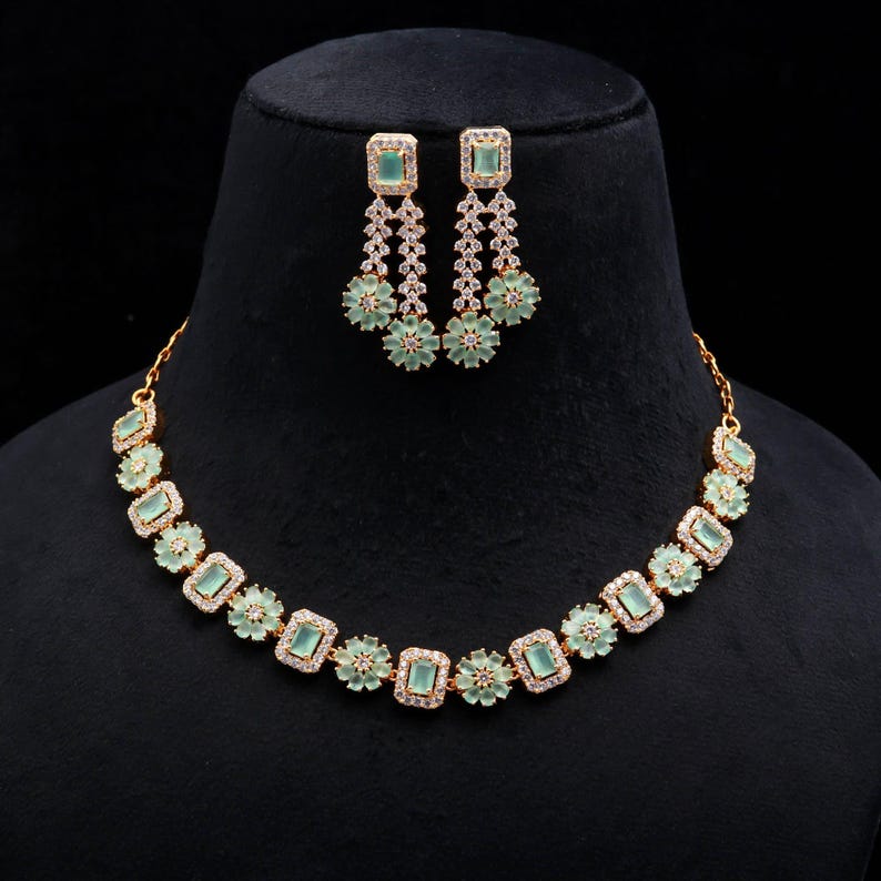 May include: A gold-toned necklace and earring set. The necklace features square, light green gemstones alternating with floral accents. The earrings have matching square gemstones and dangle with a floral design. The jewelry is displayed on a black mannequin.