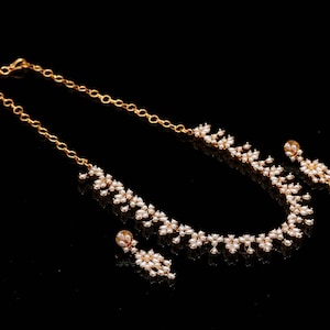 May include: A gold-colored necklace and earring set with a floral design. The necklace and earrings feature small, white, pearl-like beads. The jewelry is displayed on a black surface, creating a contrast that highlights the gold and white colors.