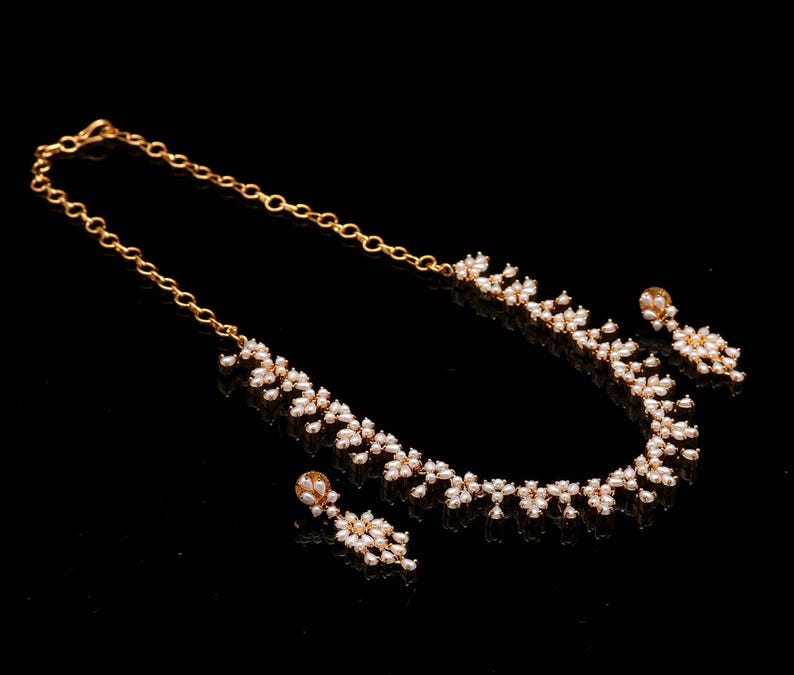 May include: A gold-colored necklace and earring set with a floral design. The necklace and earrings feature small, white, pearl-like beads. The jewelry is displayed on a black surface, creating a contrast that highlights the gold and white colors.