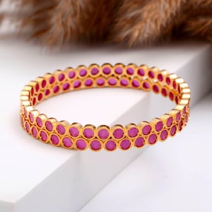 Może przedstawiać: Złota bransoletka obręczowa z dwoma rzędami żywych, różowych kamieni szlachetnych. Kamienie szlachetne są okrągłe i osadzone blisko siebie, tworząc ciągły pas koloru. Bransoletka ma polerowane wykończenie i klasyczny design.