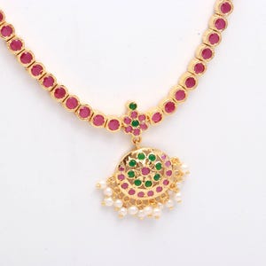 Può includere: Una collana dorata con una serie di pietre color rubino. Il ciondolo presenta un design circolare con pietre verdi e color rubino e piccole perle bianche. La collana è posta su uno sfondo bianco.