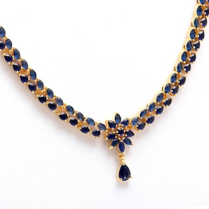 May include: A gold-toned necklace featuring a floral design with dark blue gemstones. The necklace has a central flower-shaped pendant with a teardrop-shaped gemstone dangling below. The necklace is set against a white background.