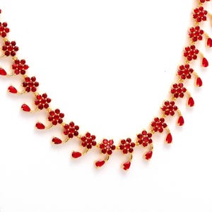 Puede incluir: Un collar dorado con un diseño floral y gemas rojas. El collar presenta racimos en forma de flor y colgantes en forma de lágrima. Las gemas son de un rojo vibrante, engastadas sobre el metal dorado, creando una pieza elegante.