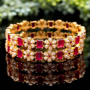 May include: Two gold bangles with a floral design. Each bangle features alternating rectangular red gemstones and white flower-shaped accents. The bangles are reflected on a dark surface.