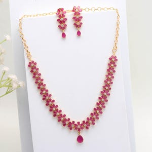 Puede incluir: Un conjunto de collar y pendientes de color dorado. El collar y los pendientes presentan un diseño floral con piedras preciosas rosas y pequeñas piedras transparentes. El collar tiene un colgante de piedra preciosa rosa en forma de lágrima. Los pendientes tienen una piedra preciosa rosa.