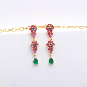 May include: A pair of gold-toned dangle earrings. Each earring features a cluster of small, pink gemstones with a central green gemstone. A teardrop-shaped green gemstone hangs from each earring.